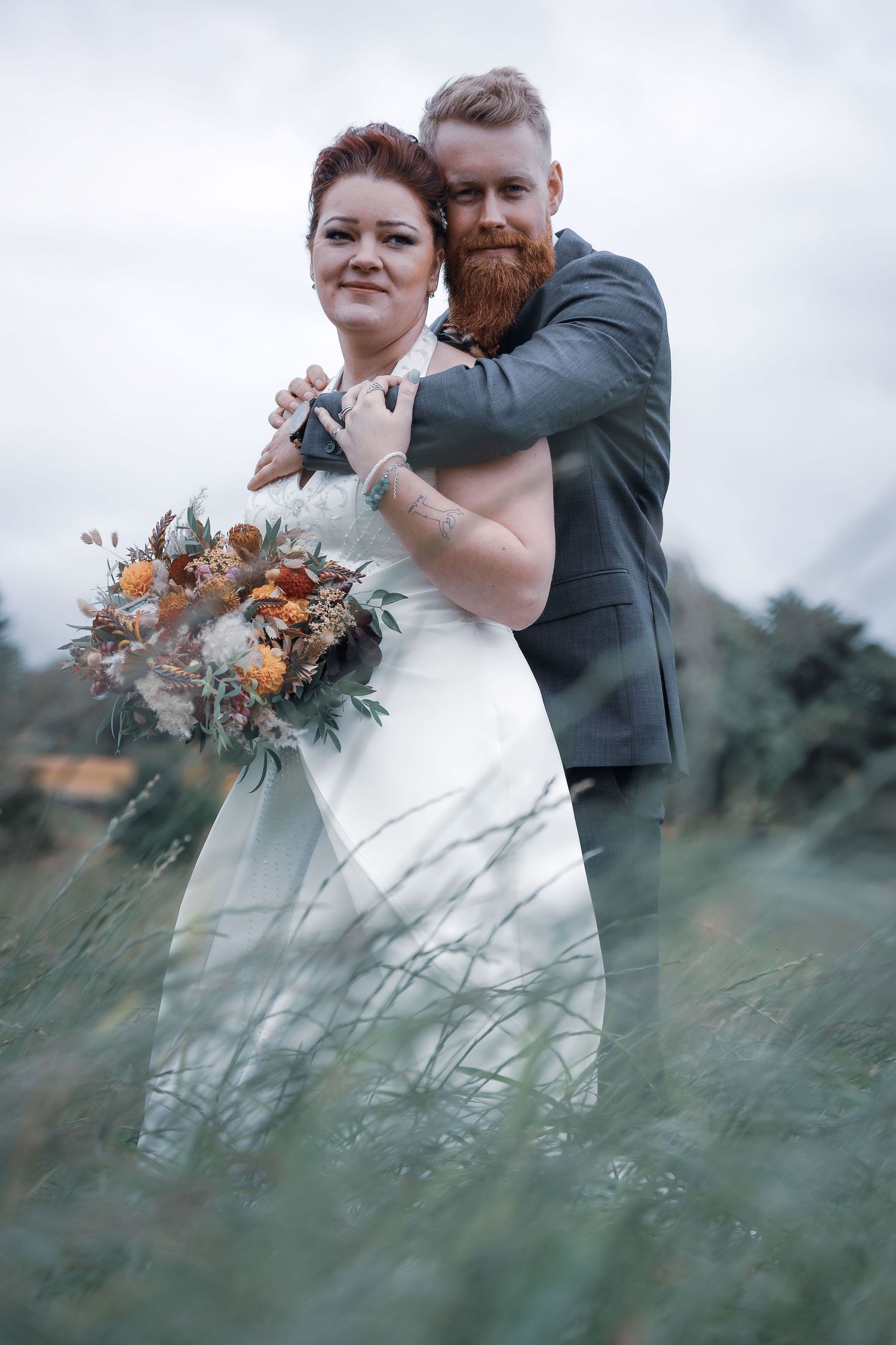 Bearded groom embracing bride holding autumn bouquet in tall grass field.