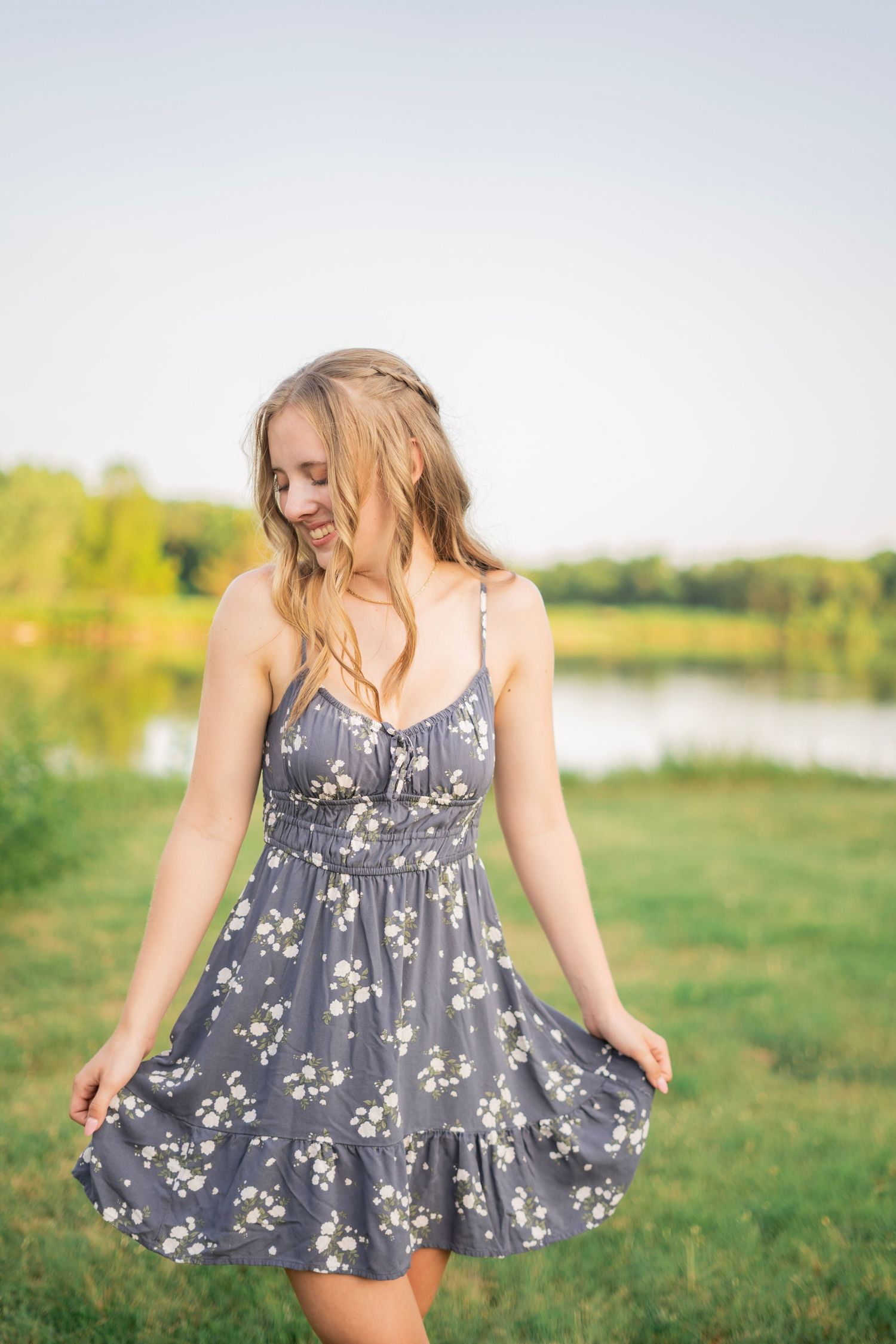 A young blonde woman in a gray floral sundress stands near a lake in a sunny green field.