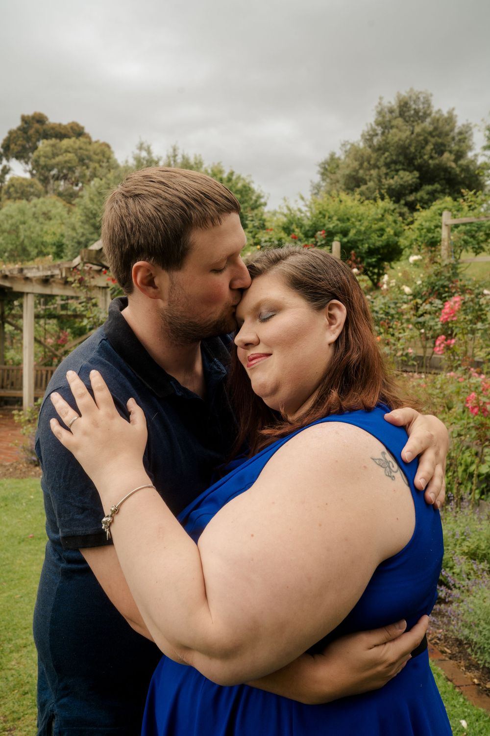 casually dressed man kisses plus size fiance dressed in blue on forehead
