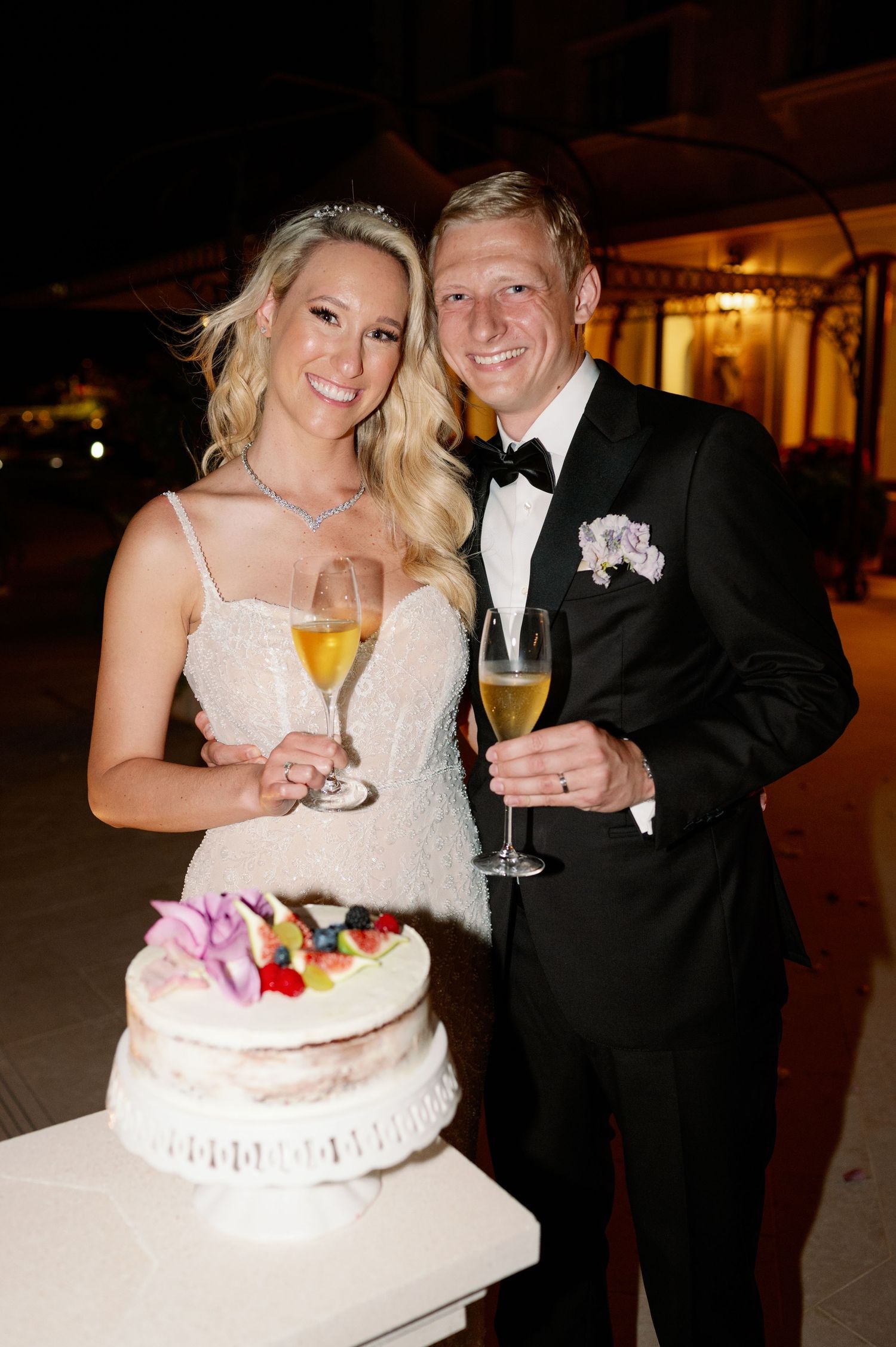 Happy couple poses with champagne next to their wedding cake at evening reception.