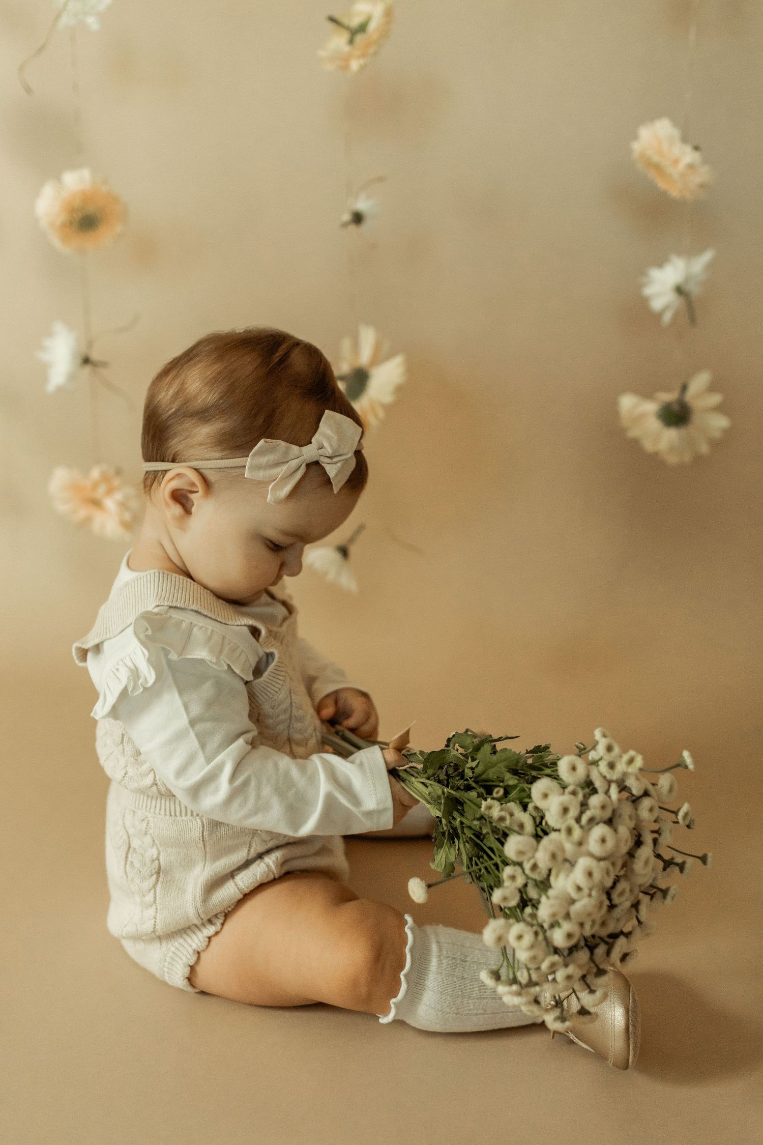 Baby in white outfit sits with floral decorations and holds delicate white flowers against beige backdrop.