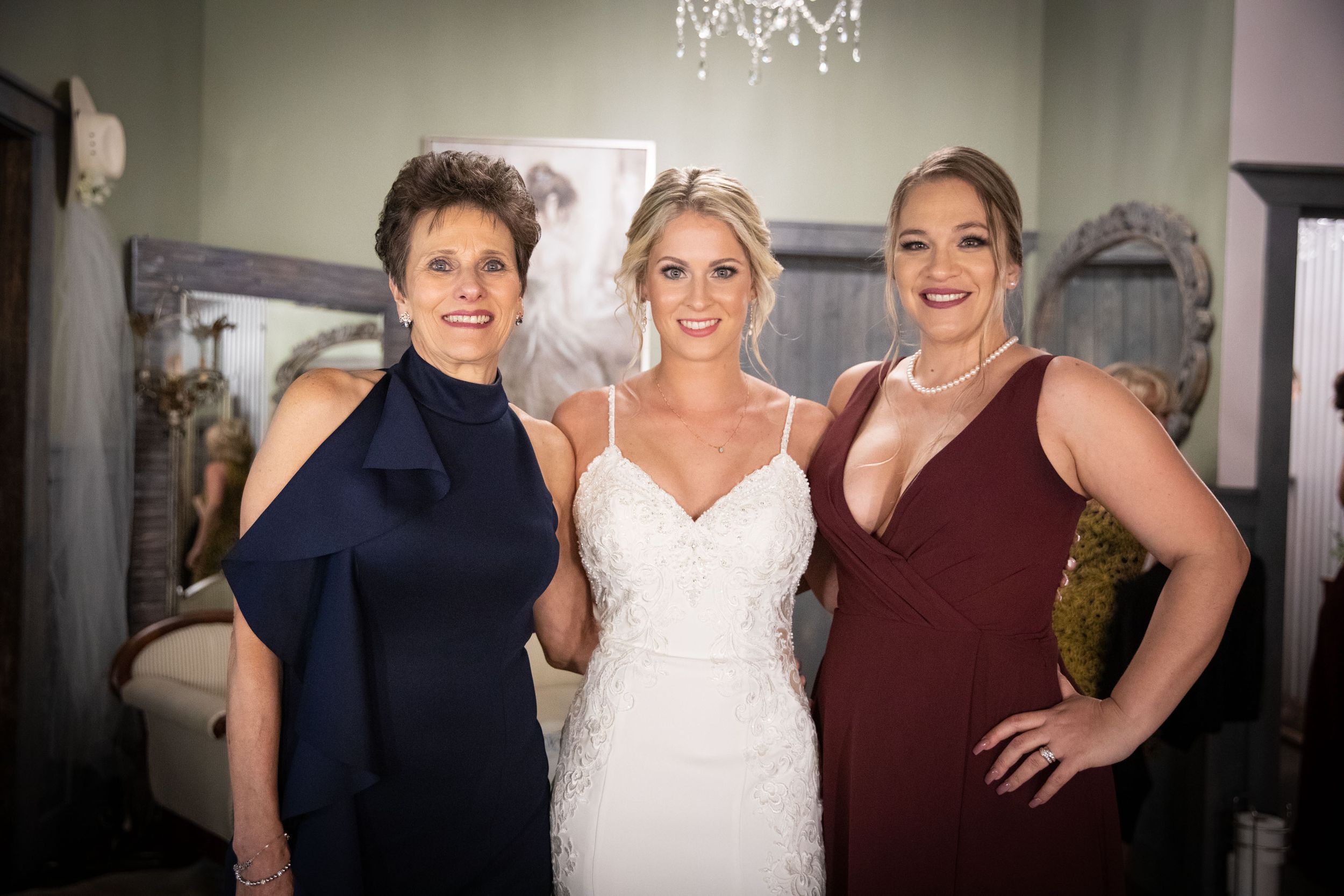 Three women pose together in elegant evening wear under a crystal chandelier.