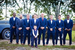 Wedding party in matching navy suits poses with vintage car at outdoor venue.