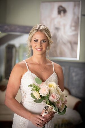 Bride in white spaghetti strap dress holds pink rose bouquet during wedding preparations.