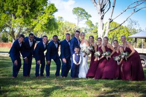 Wedding party in navy suits and burgundy dresses poses on grassy lawn.