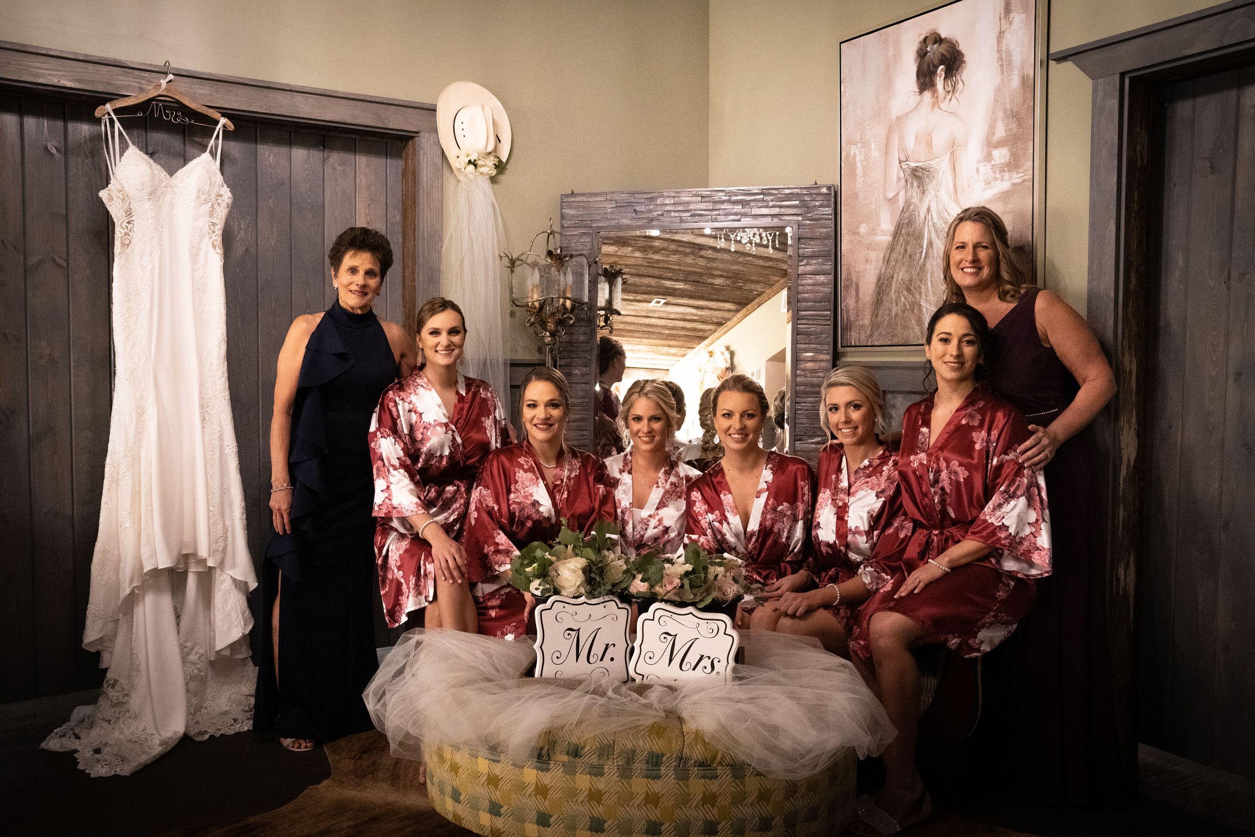 Wedding dress hangs while bridal party in matching robes gathers in preparation room.