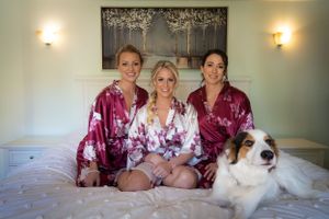 Bridal party in burgundy robes poses on bed with white dog.