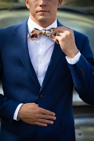 Close-up of groom adjusting floral bow tie while wearing navy blue suit.