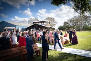 Outdoor wedding ceremony guests seated on wooden benches watching couple on white aisle runner.