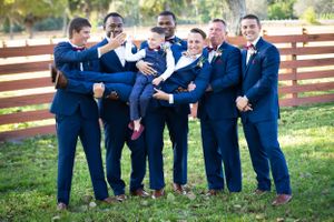 Groomsmen in navy blue suits pose playfully for a group photo outdoors by a wooden fence.