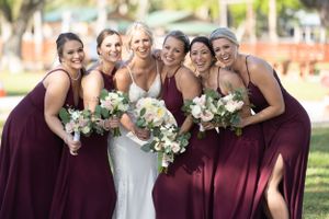 Wedding party in burgundy dresses and white gown pose with bouquets against sunny outdoor backdrop.