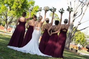 Wedding party raises floral bouquets while posing with backs to camera in burgundy dresses and white gown.
