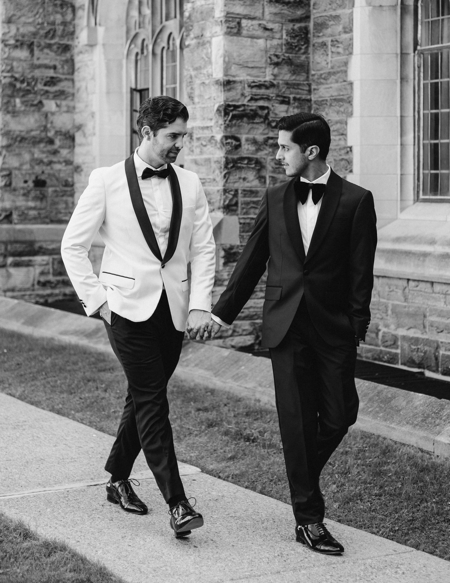 Two grooms in formal wear walking hand in hand along a stone building pathway.