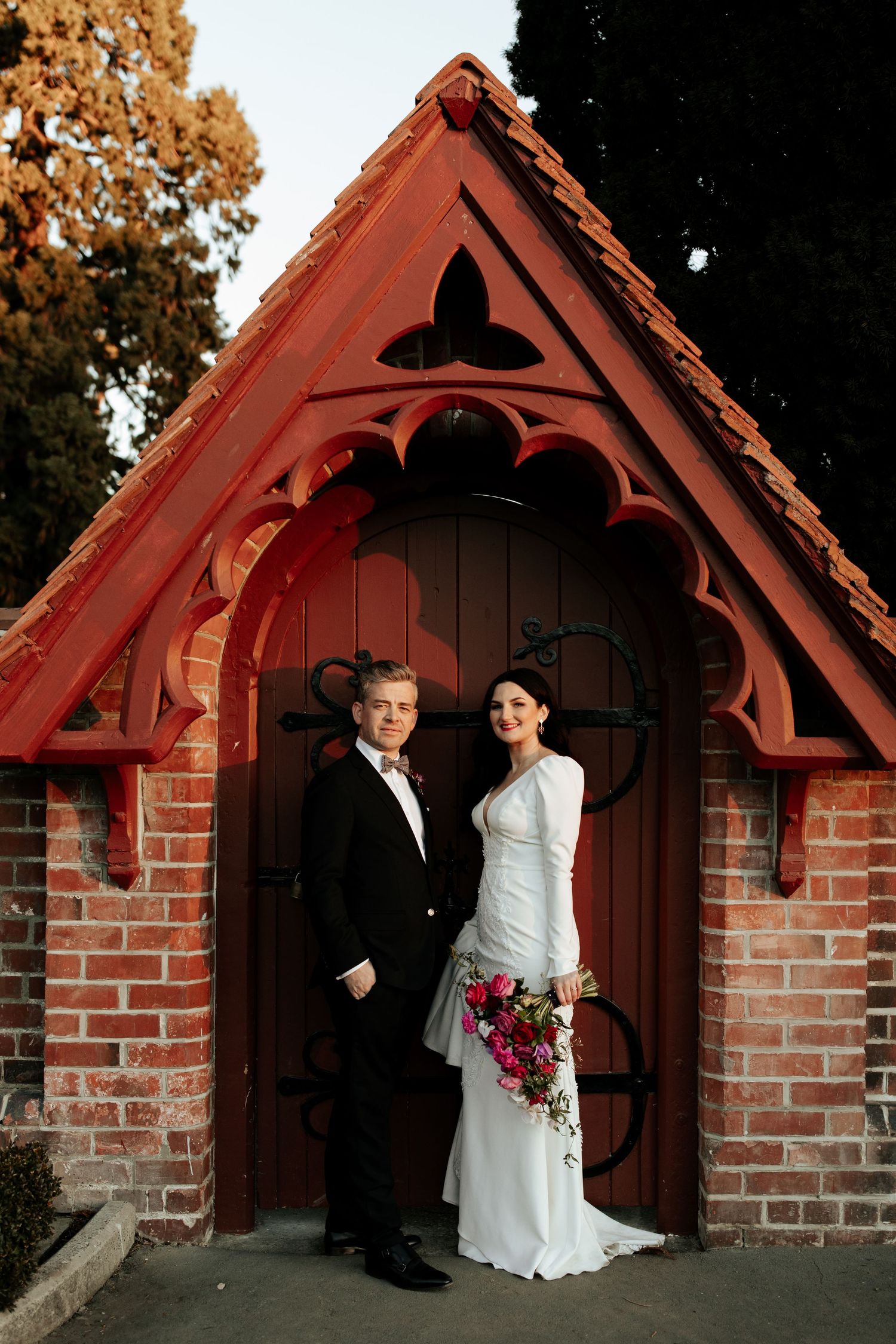 A couple stands in front of a red Victorian Gothic-style doorway with ornate woodwork.