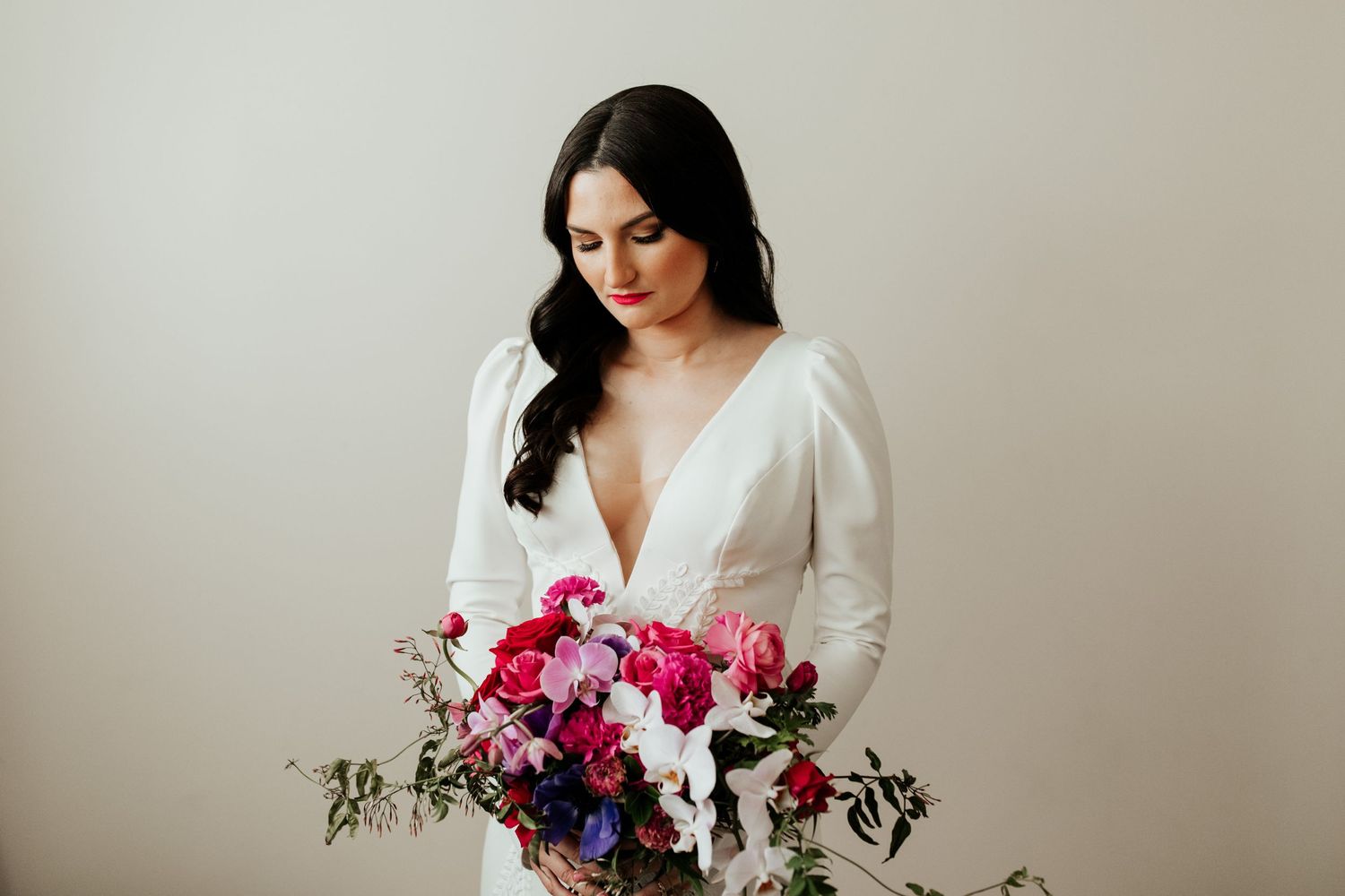 Elegant white wedding gown with deep V neckline and vibrant pink and purple floral bouquet against neutral background.