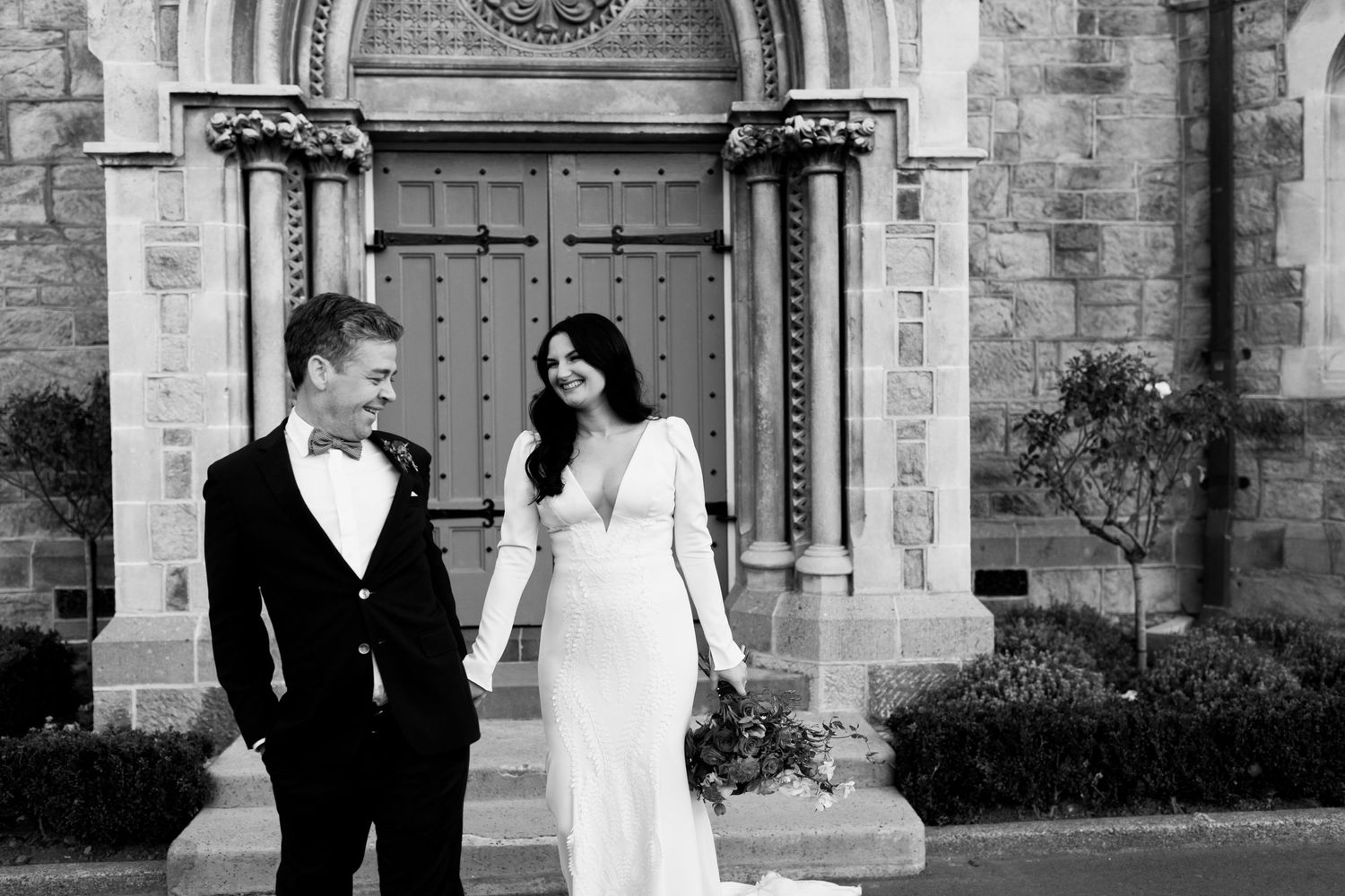 Black and white portrait of wedding couple standing in front of historic church entrance.