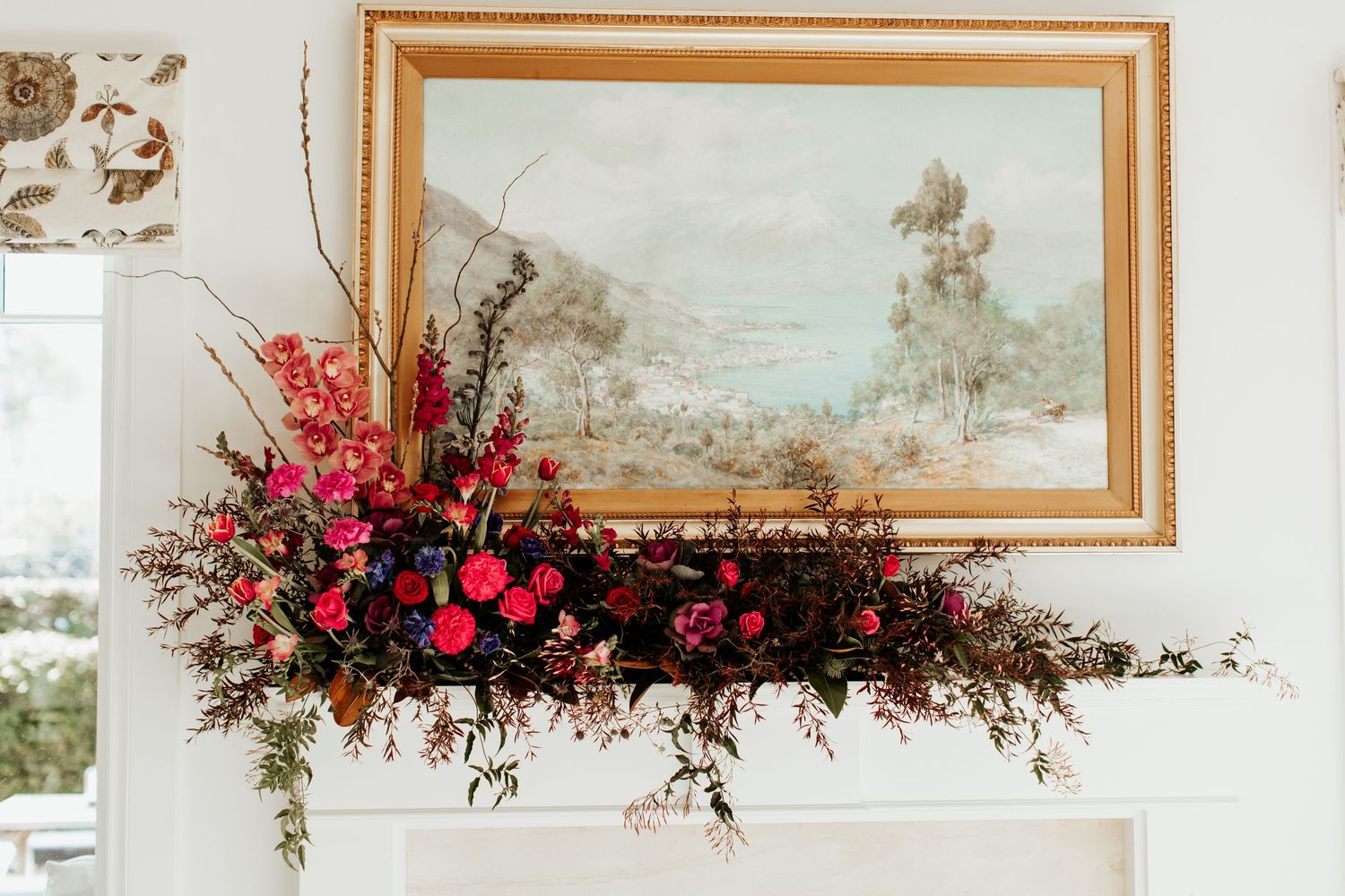 Large floral arrangement in red and pink tones decorating mantel beneath framed landscape artwork.
