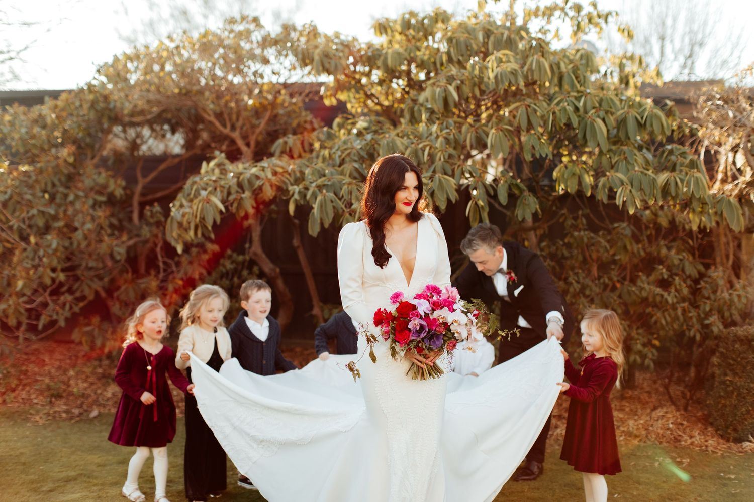 A scenic outdoor wedding photo features children in burgundy attire surrounding a flowing white dress.