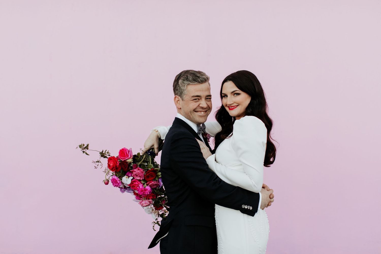 Romantic couple embrace with colorful floral bouquet against pink wall.