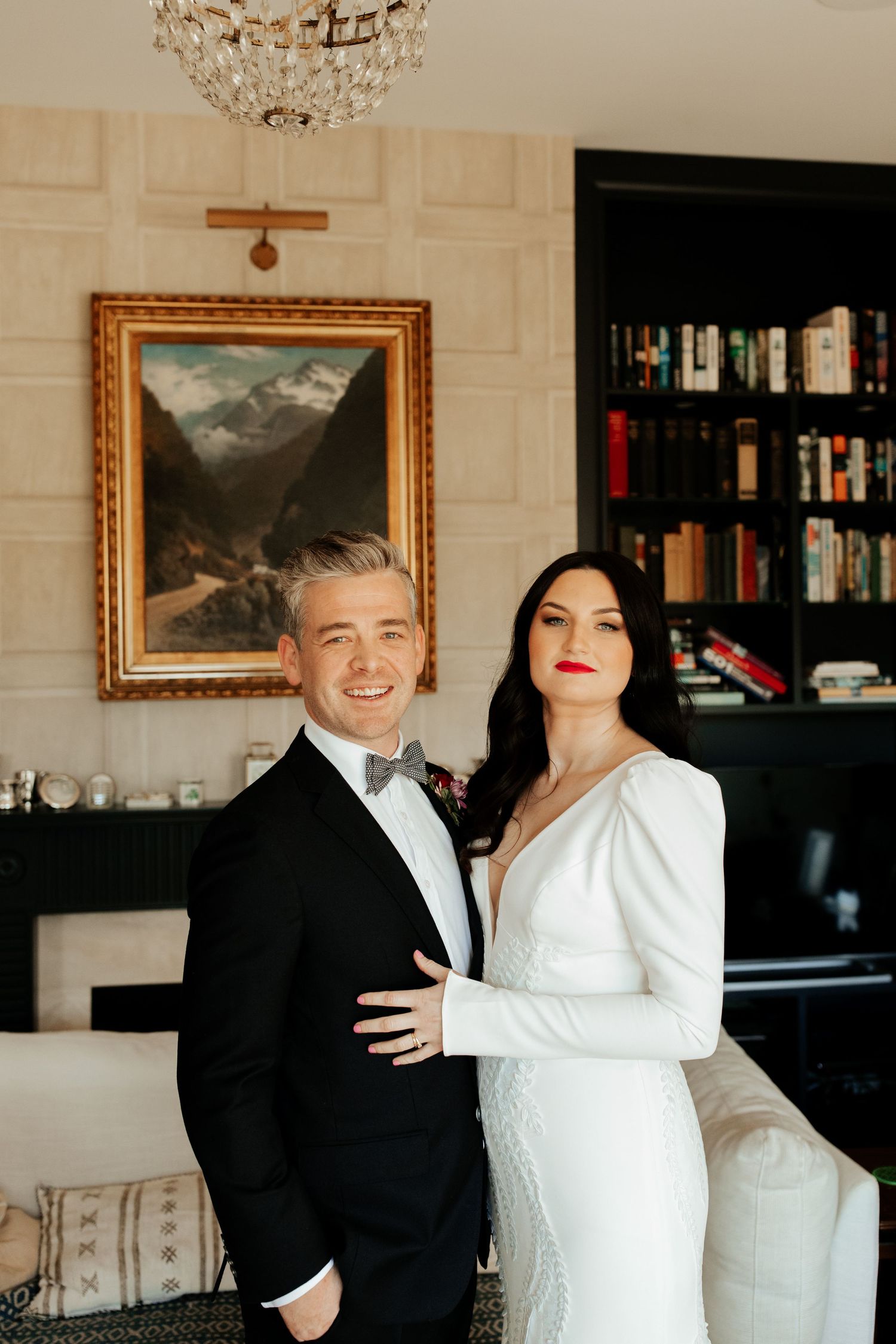 Formal portrait of a couple in wedding attire standing in an elegant room with built-in bookshelves.