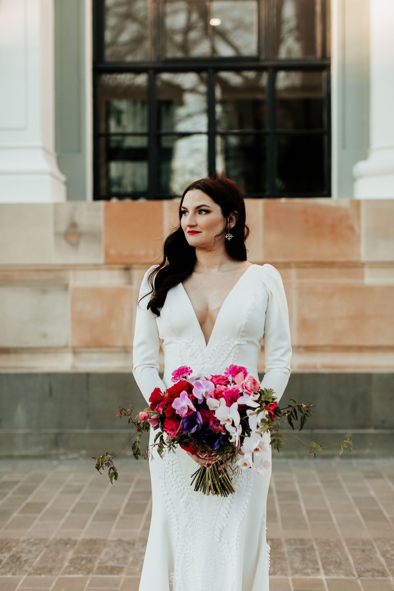 Bride with long dark hair holding vibrant pink and purple bouquet against stone building.