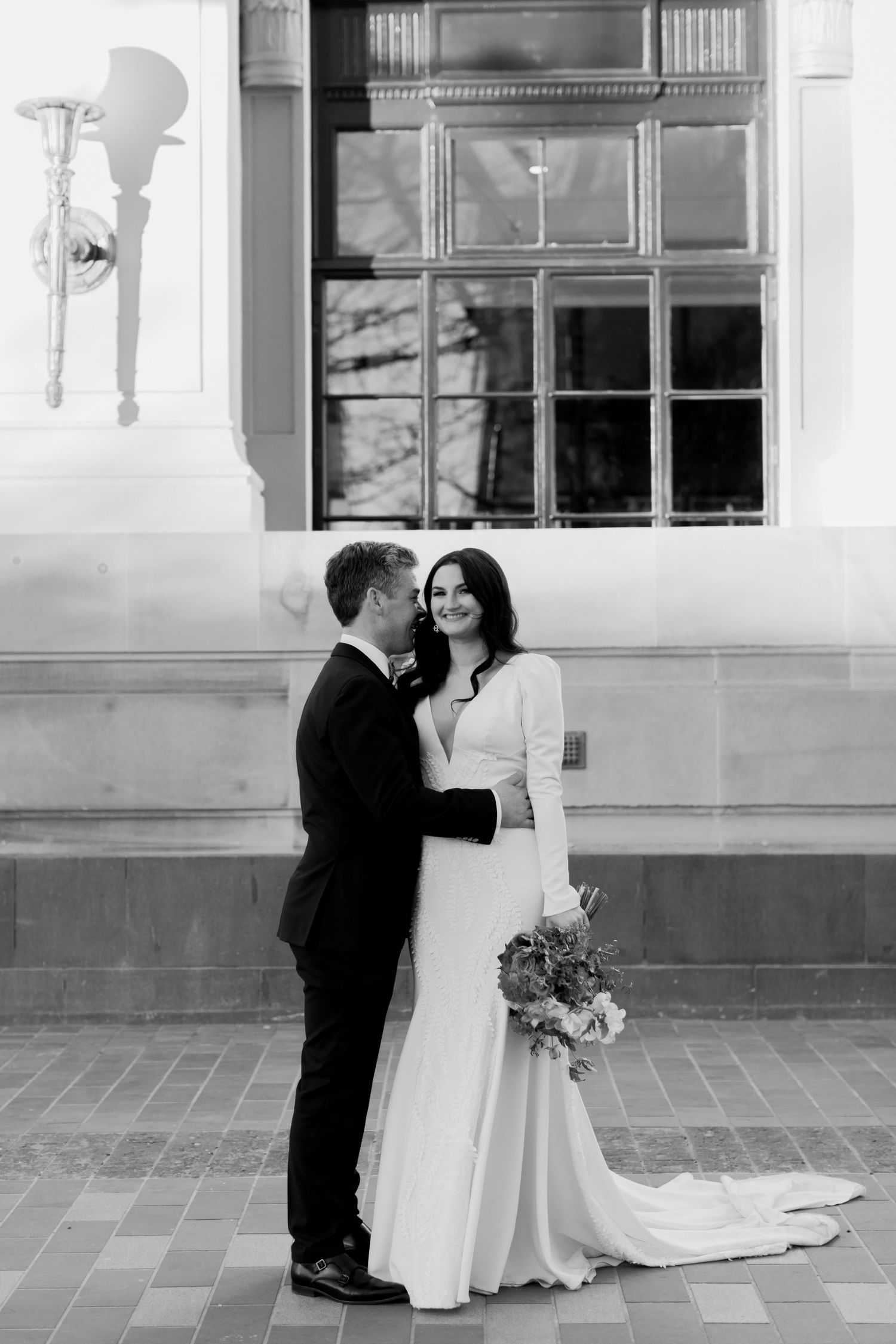 Black and white portrait of newlyweds embracing against architectural backdrop.