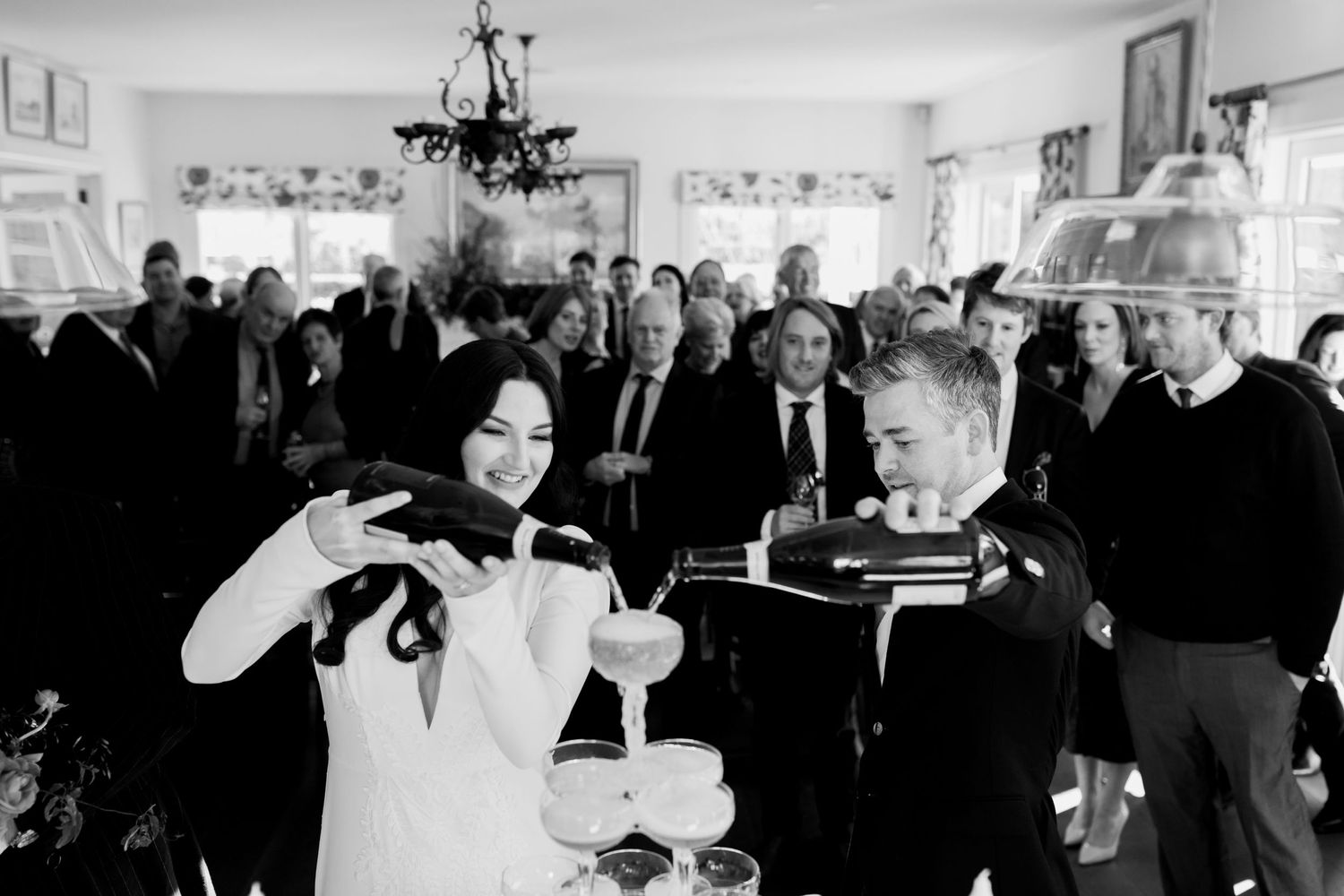 A black and white photo captures guests pouring champagne into stacked glasses at a wedding reception.