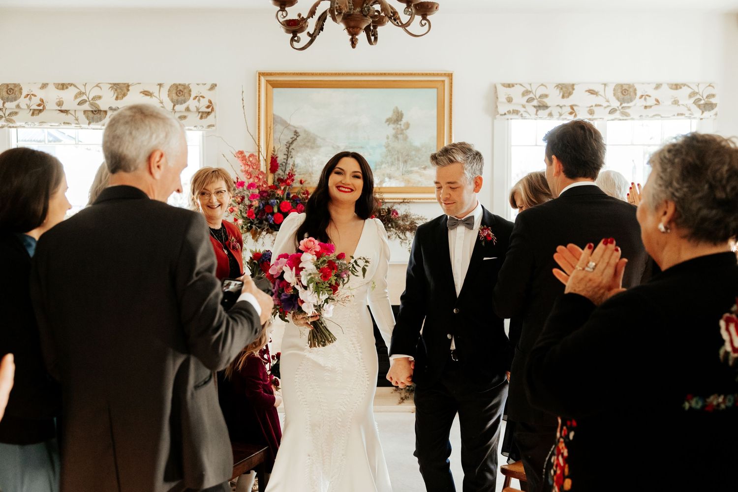A wedding celebration shows guests applauding as a couple walks through an elegant room with floral decor.