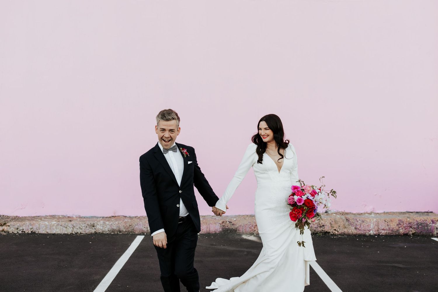 Newlyweds share joyful moment in parking lot with pink wall backdrop and floral bouquet.