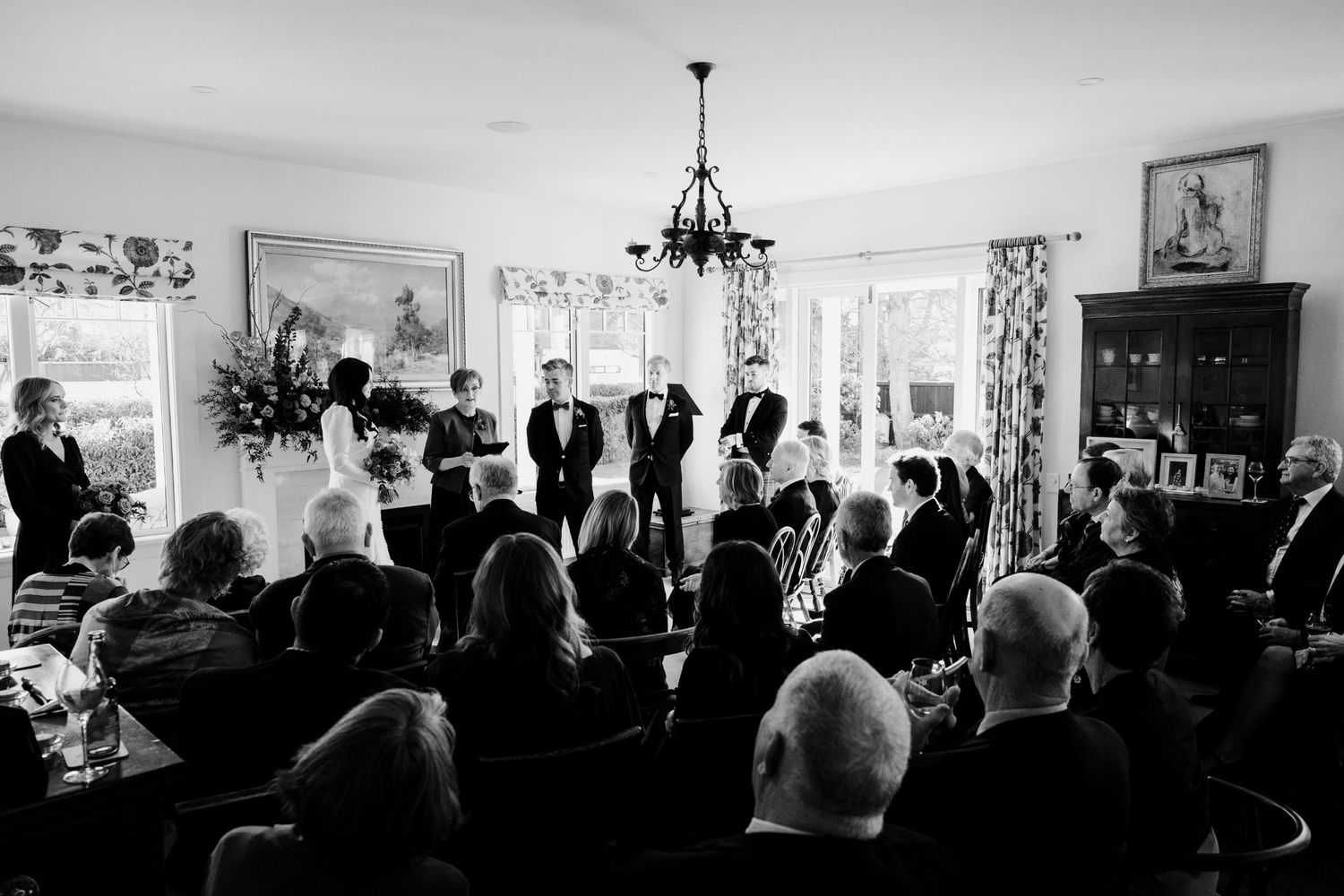 A black and white photo of an intimate indoor wedding ceremony with guests gathered in a bright room.