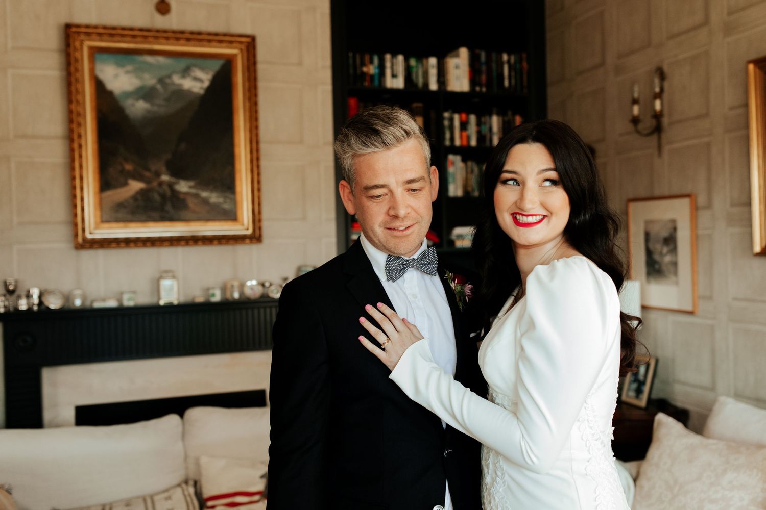Elegant couple posing in formal attire in front of a fireplace with landscape painting.