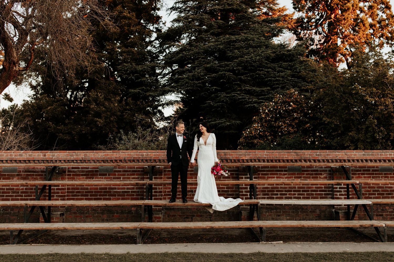 A couple in wedding attire stands on tiered brick steps with evergreen trees in the background.