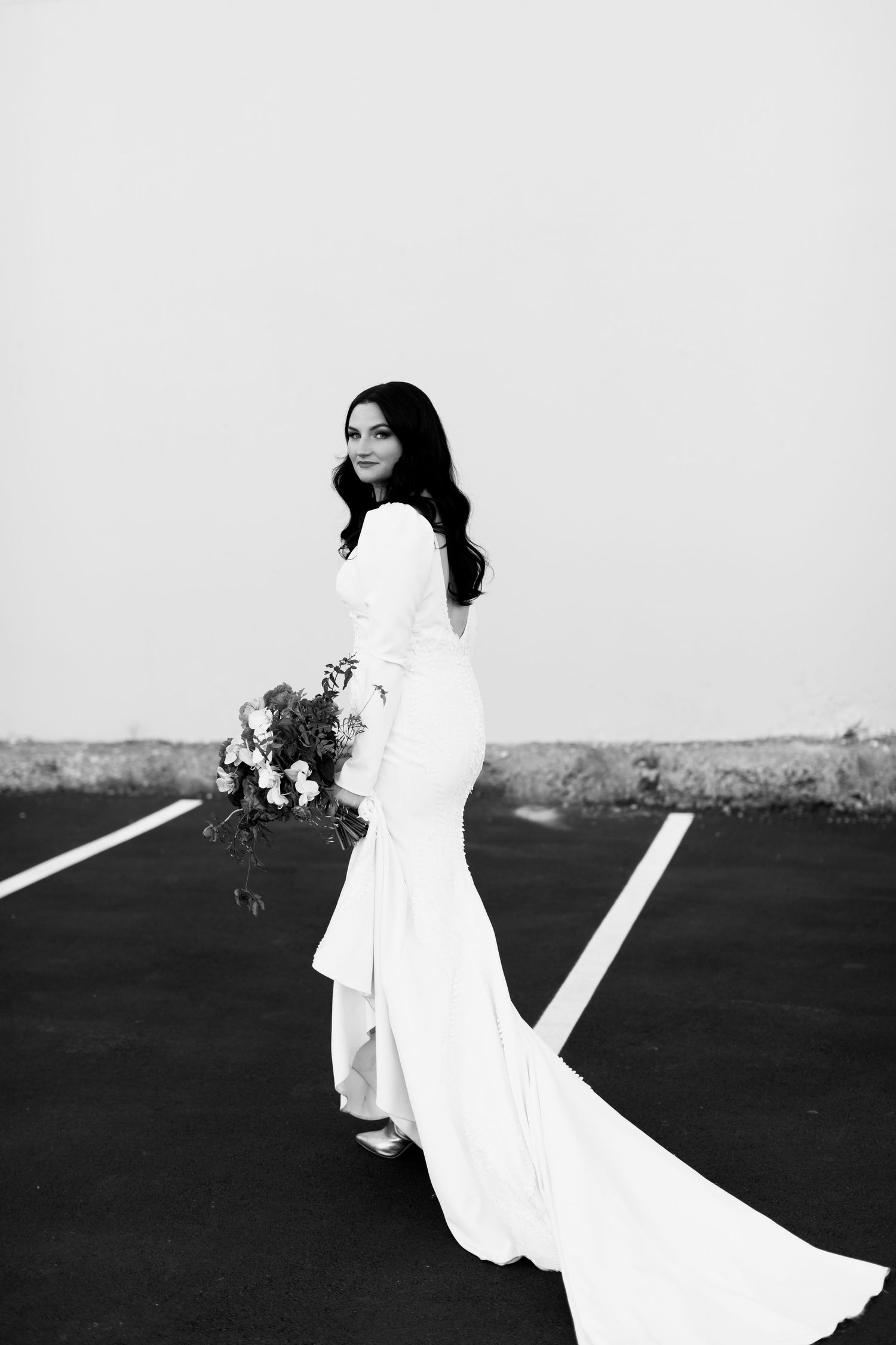 Elegant bride in fitted white gown poses with bouquet against minimal backdrop.