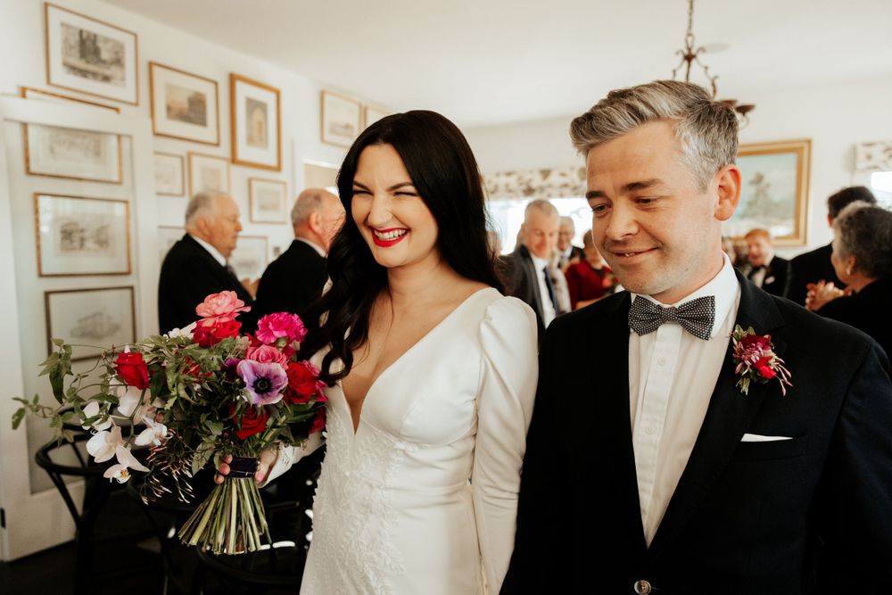 A couple smiles joyfully at their wedding while holding a vibrant pink and red bouquet.