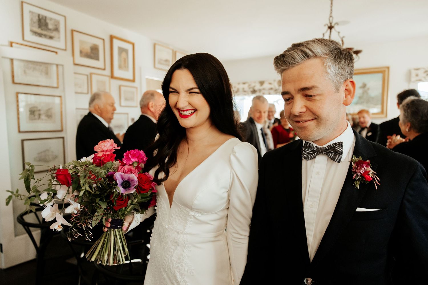 A couple smiles joyfully at their wedding while holding a vibrant pink and red bouquet.