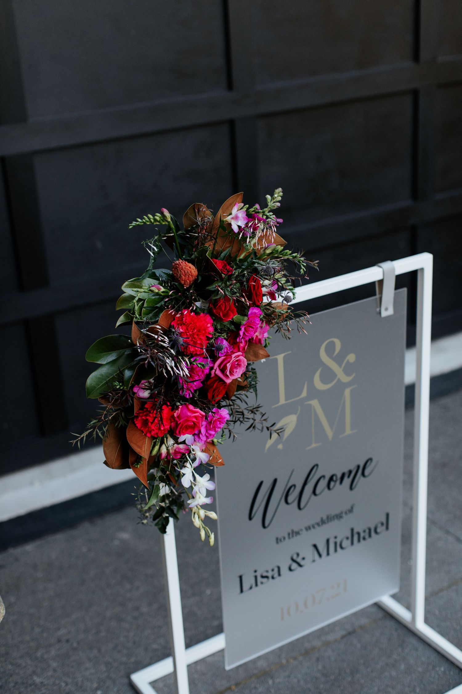 Modern wedding welcome sign decorated with vibrant red and pink florals against a dark background.