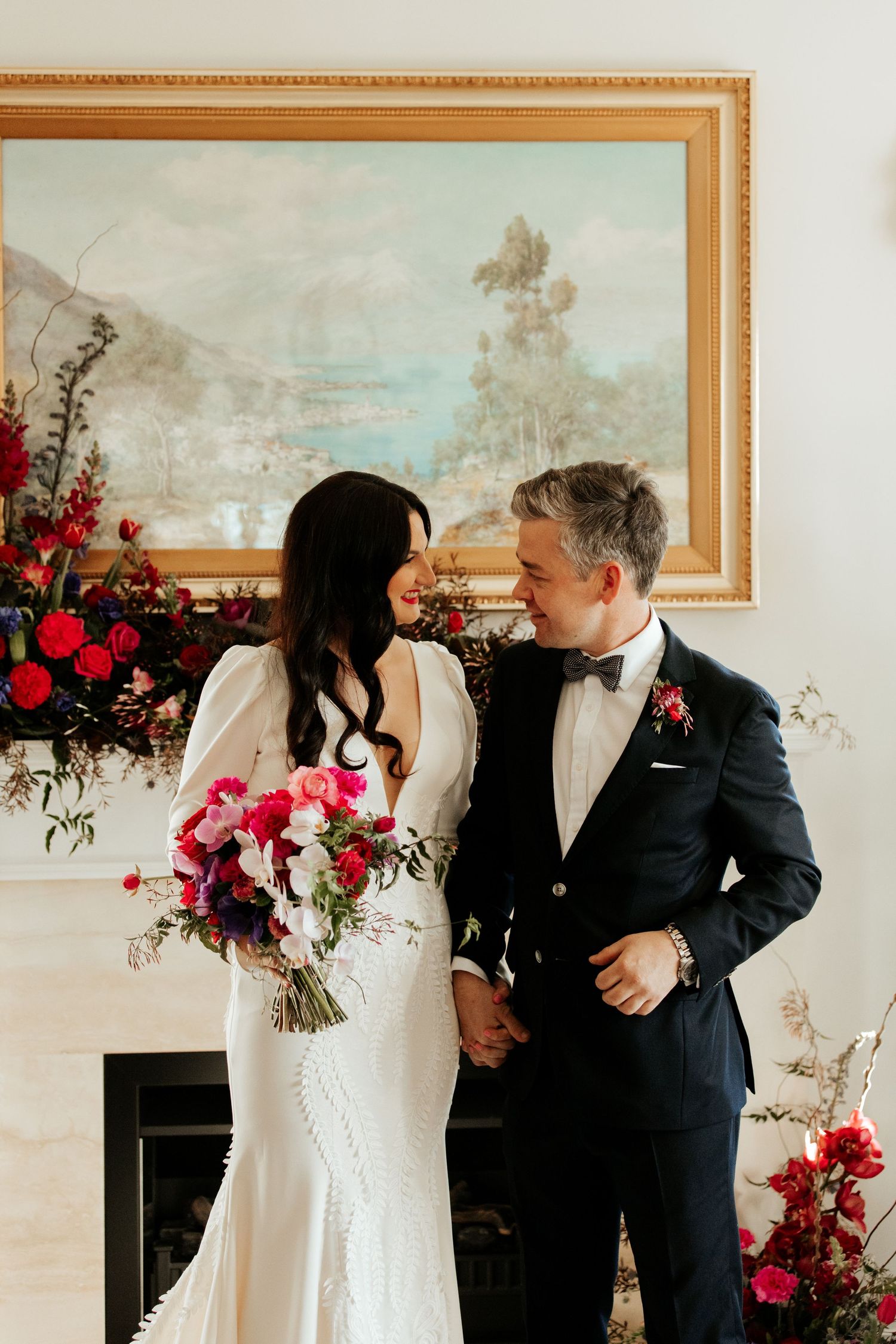 Couple in wedding attire posing by a fireplace decorated with vibrant red and pink flowers.