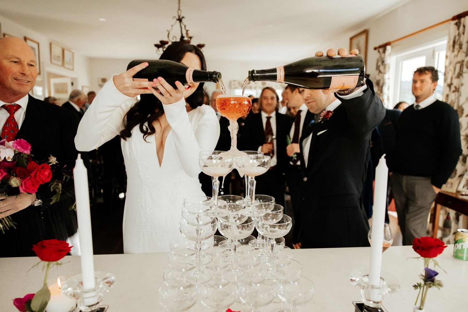Wedding guests participate in a champagne tower pouring celebration indoors.