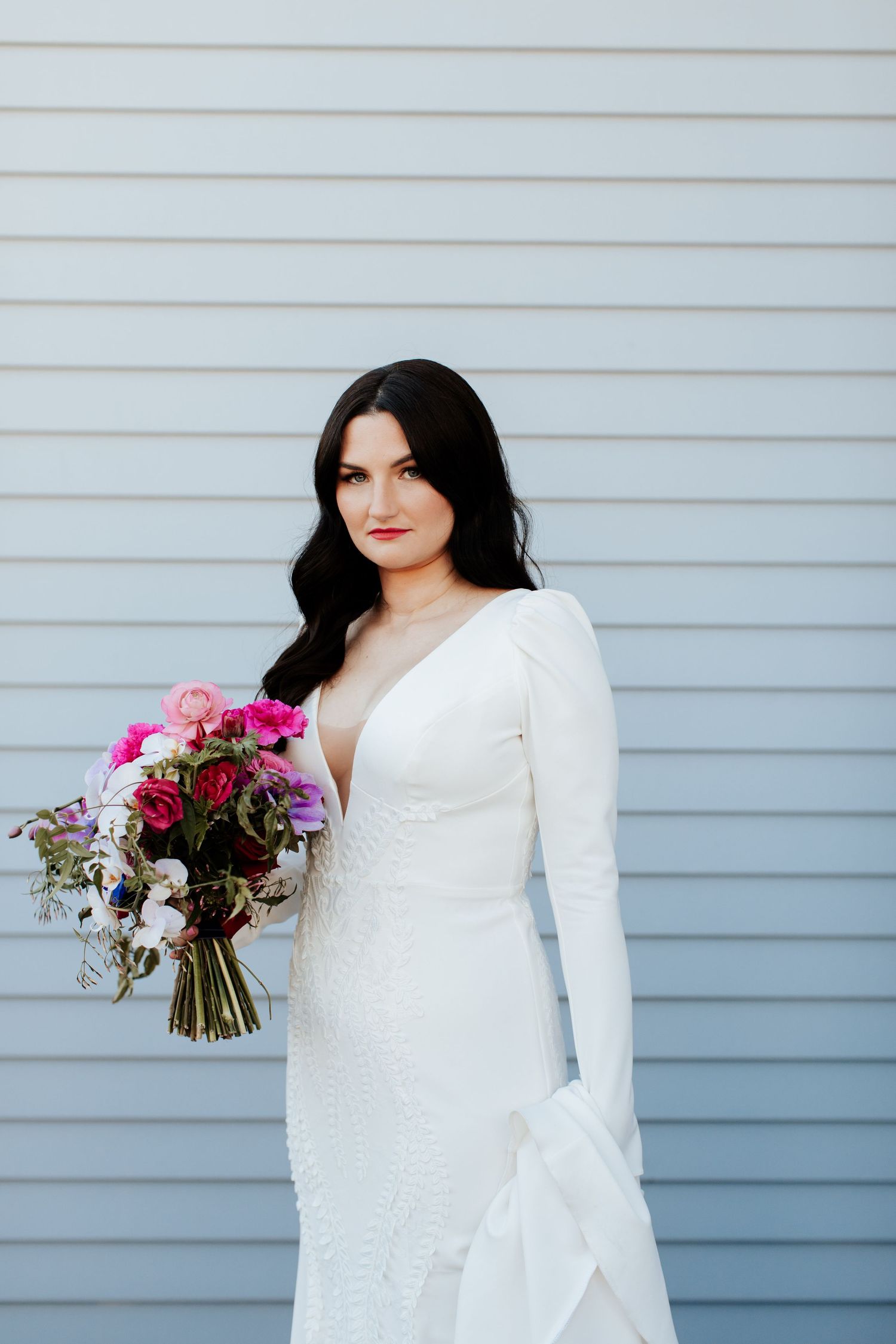 Modern bridal portrait against grey siding with fitted white gown and colorful bouquet.