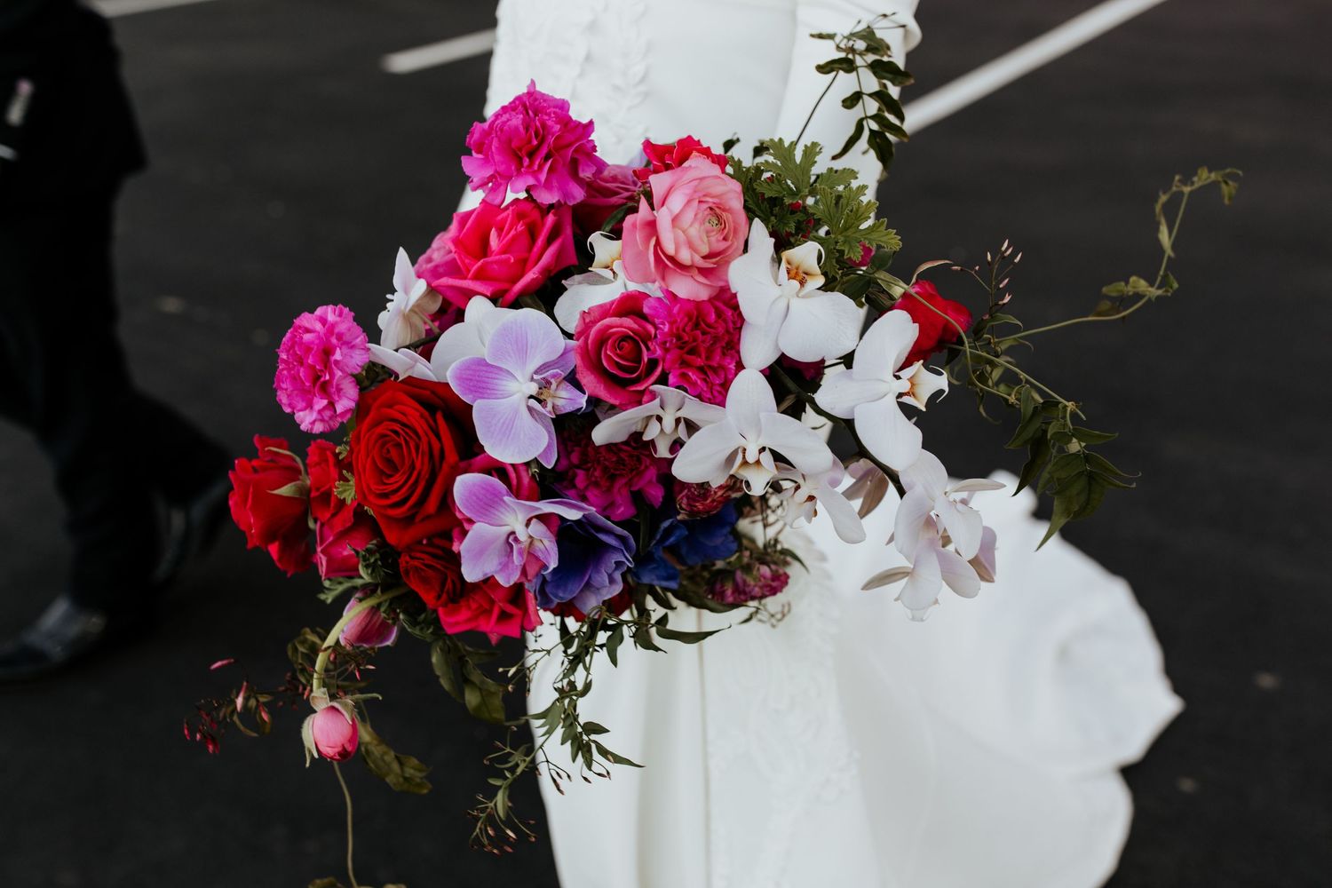 Vibrant wedding bouquet with pink and red flowers against white dress detail.