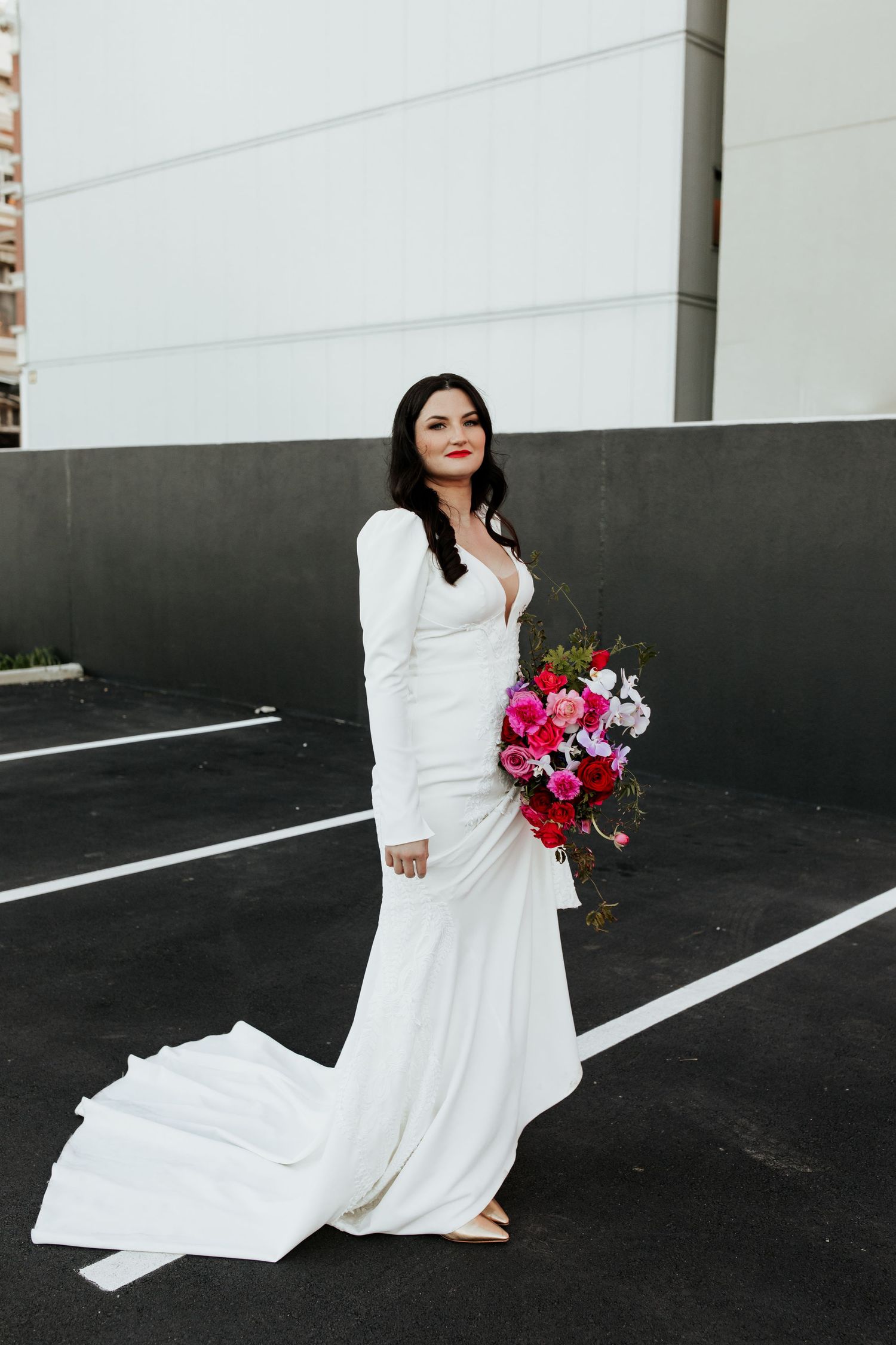 Bride in long-sleeved white gown poses with colorful bouquet in urban setting.