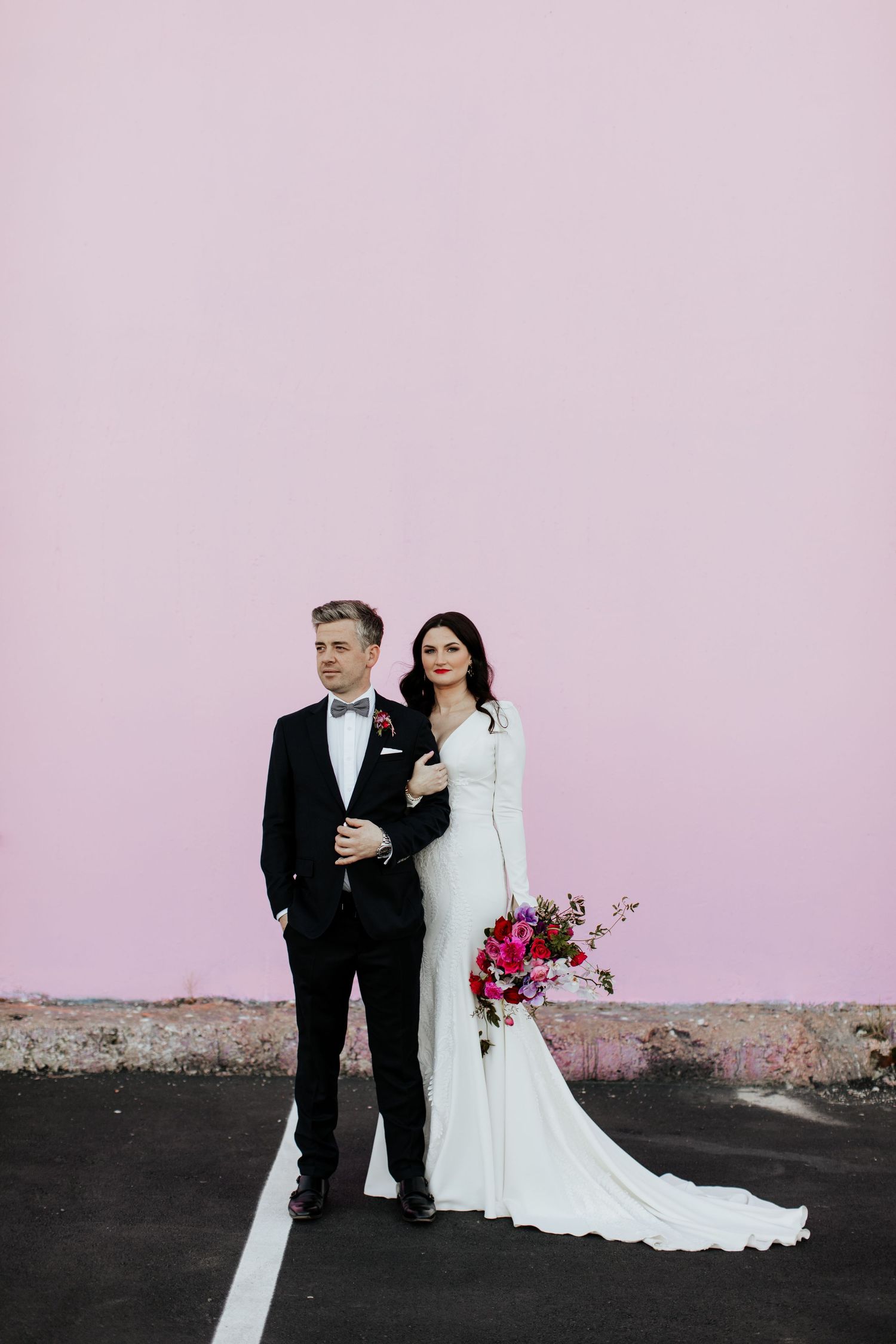 Stylish wedding couple posing against pink wall with bright floral bouquet.