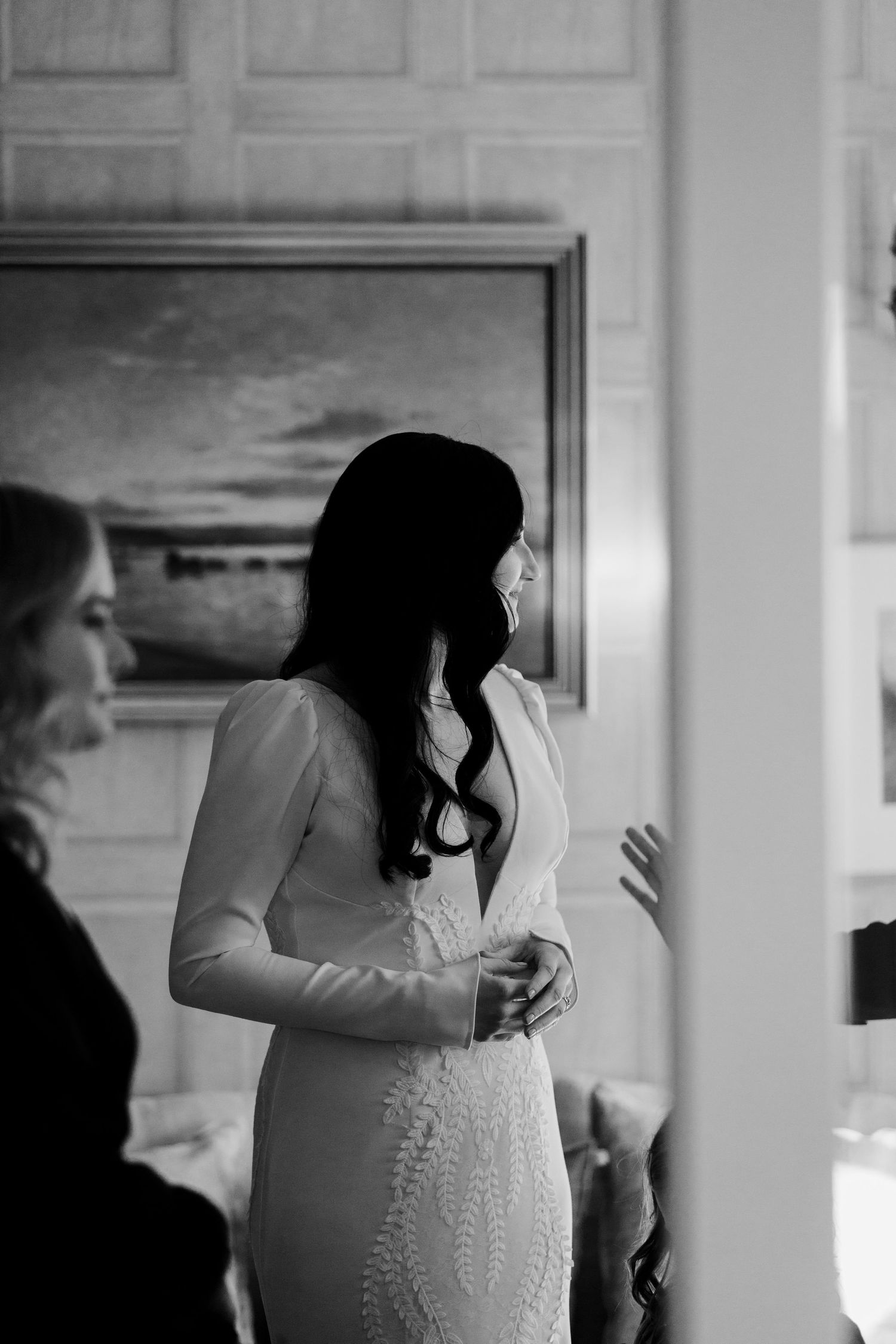 Black and white portrait of a bride in a long-sleeved white gown standing in an elegant interior.