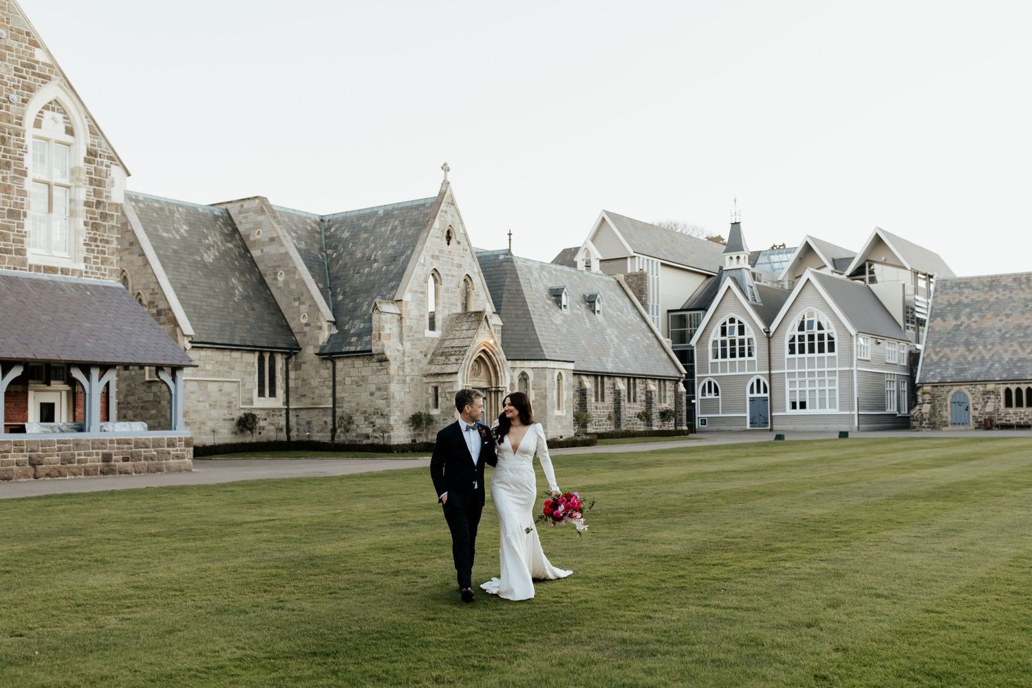 Newlyweds walking on grass in front of historic stone buildings with gothic architecture.
