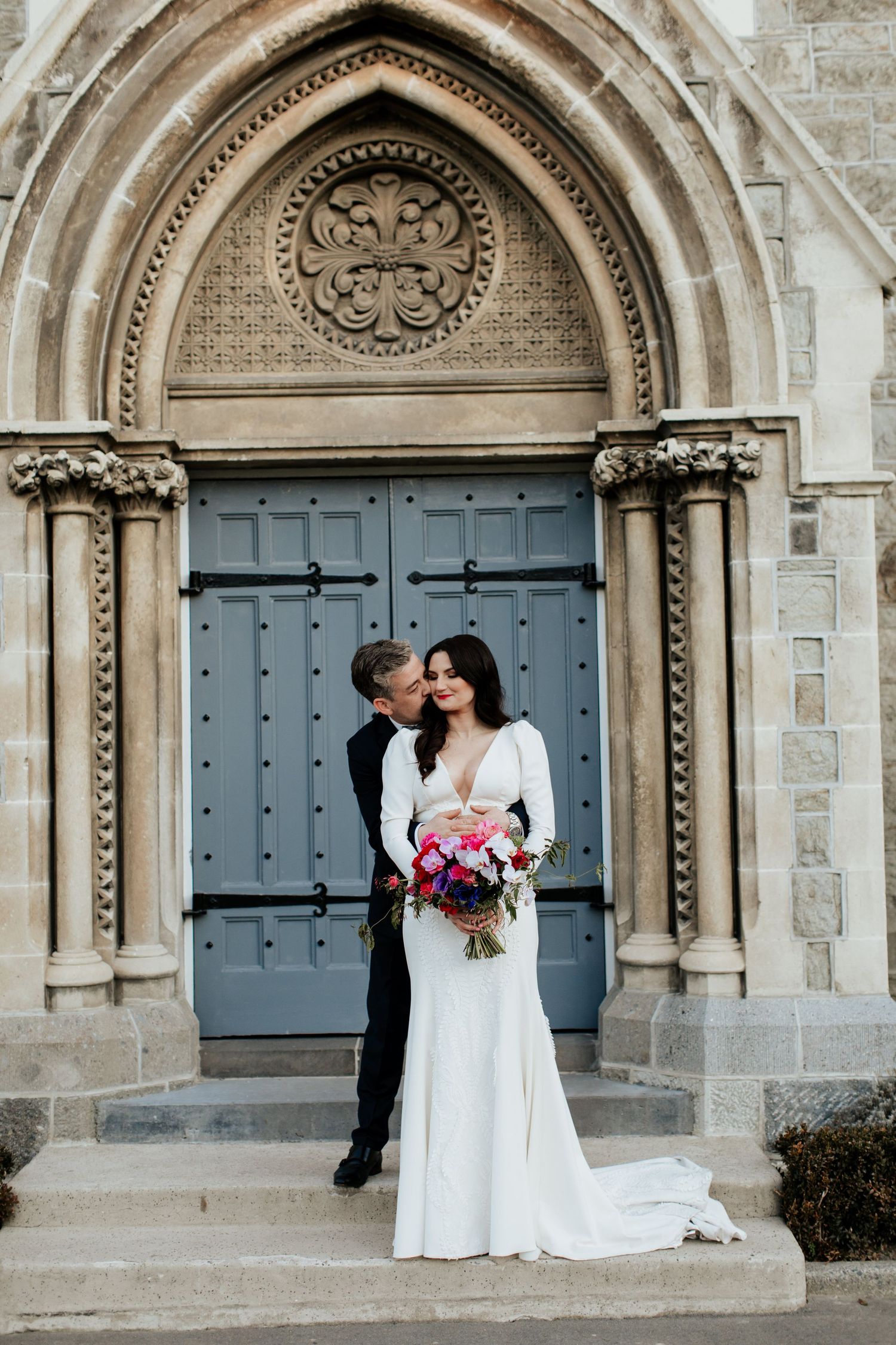 A couple shares an intimate moment in front of an ornate Gothic church doorway with stone arches.