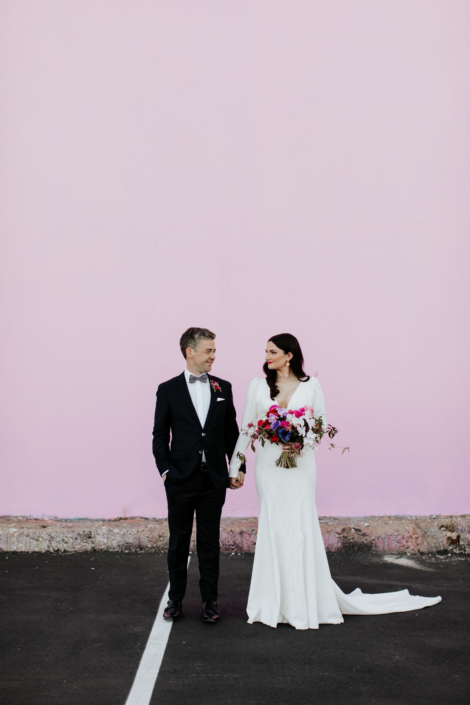 Wedding couple poses against pink wall in formal attire with bright floral bouquet.