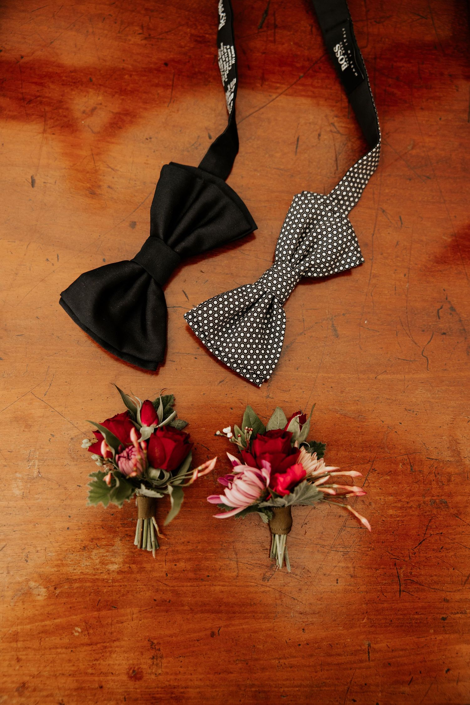 Black and polka dot bow ties with small red flower boutonnieres on wooden surface.