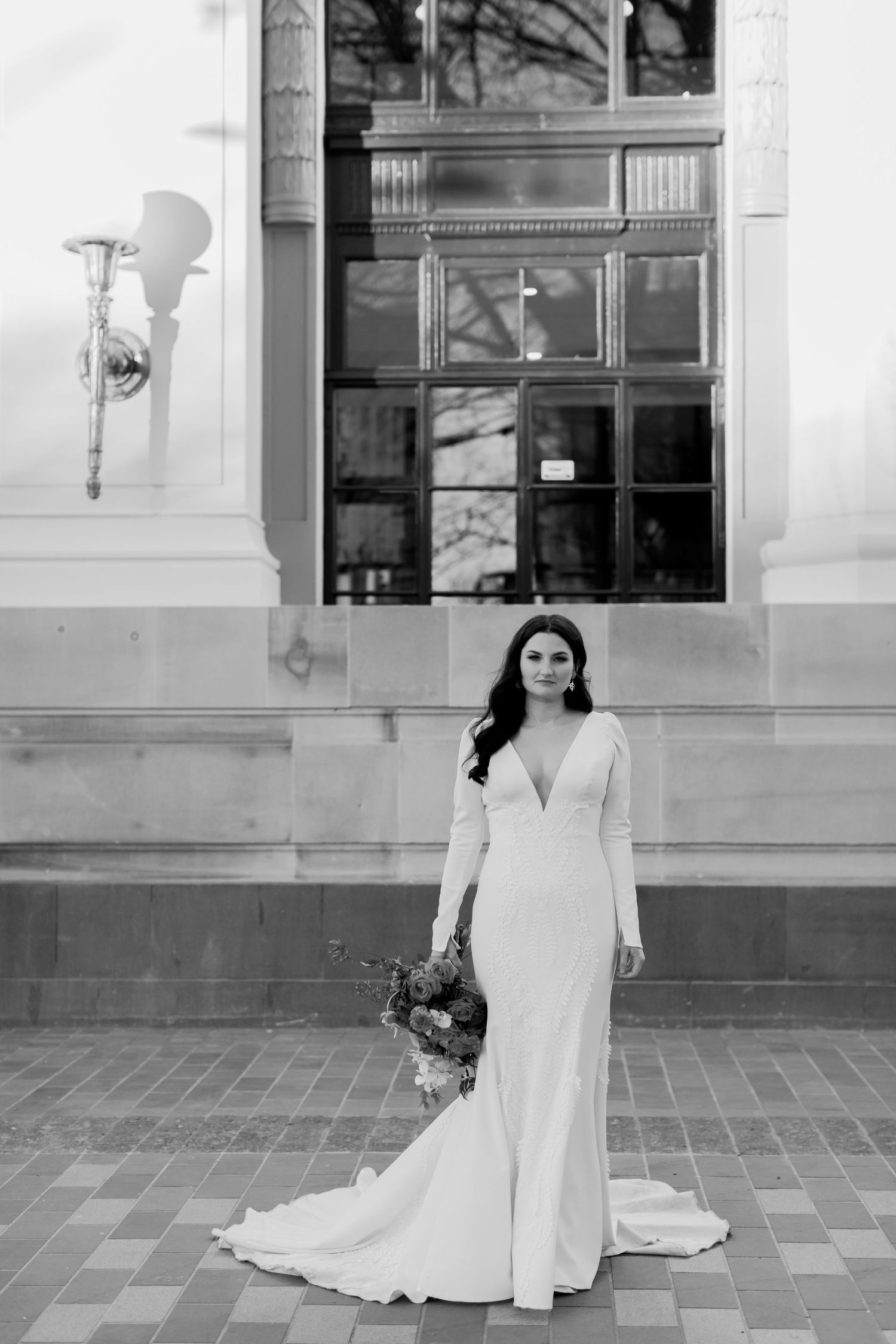 Monochrome bridal portrait with elegant white dress and vibrant bouquet against architectural setting.