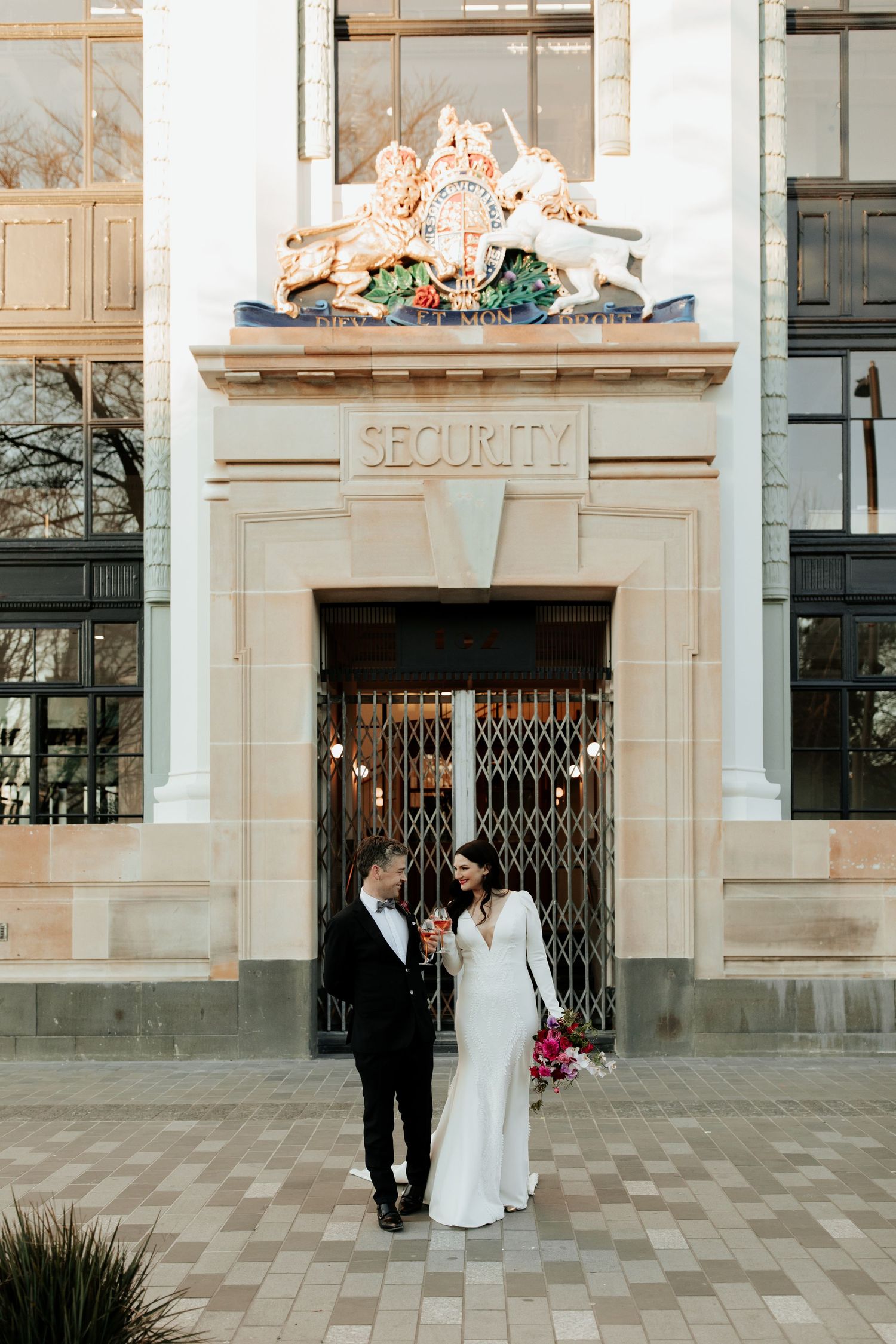 A couple poses beneath an ornate security entrance with decorative stonework and a floral arrangement.