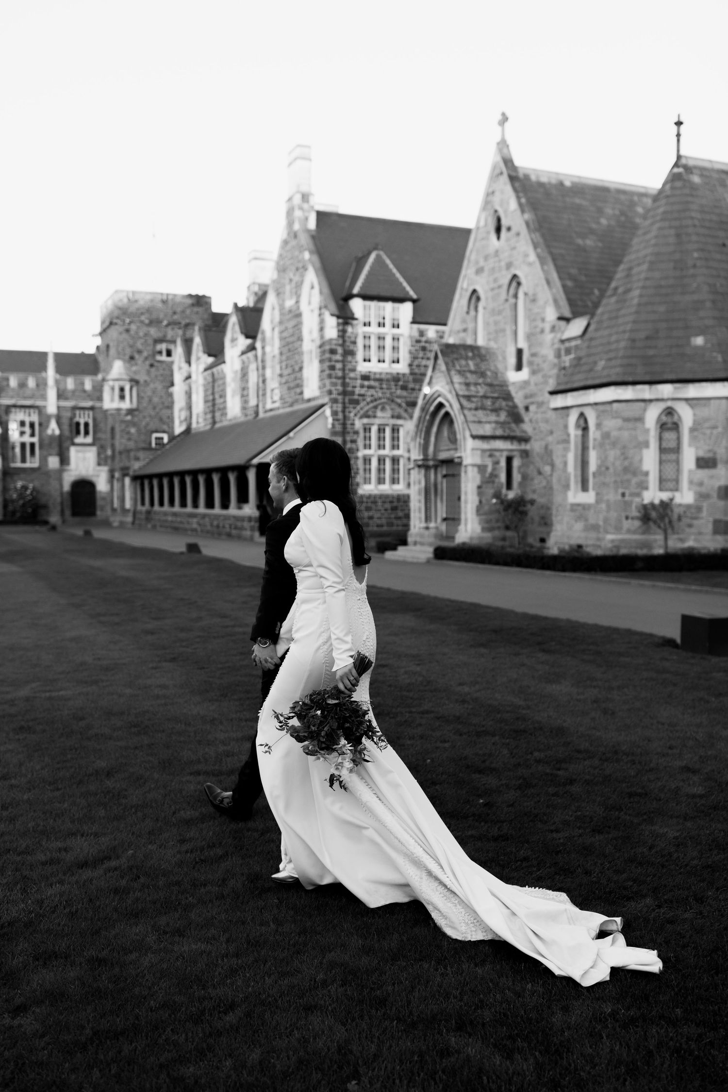 A flowing white dress trails behind on grass near Gothic revival architecture in black and white.